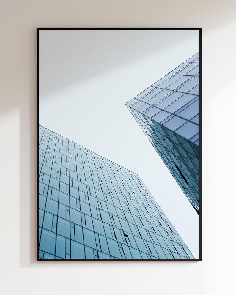 May include: Framed architectural print featuring two modern buildings with blue-tinted glass facades against a white sky. The buildings have a geometric design with numerous windows, creating a contemporary aesthetic.