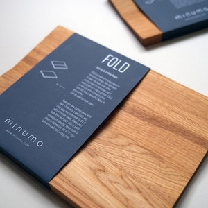 May include: Two rectangular wooden cutting boards with a natural grain finish. Each board features a dark blue paper band wrapped around one end, printed with the white text FOLD, Minumo, and care instructions for the kitchen product.