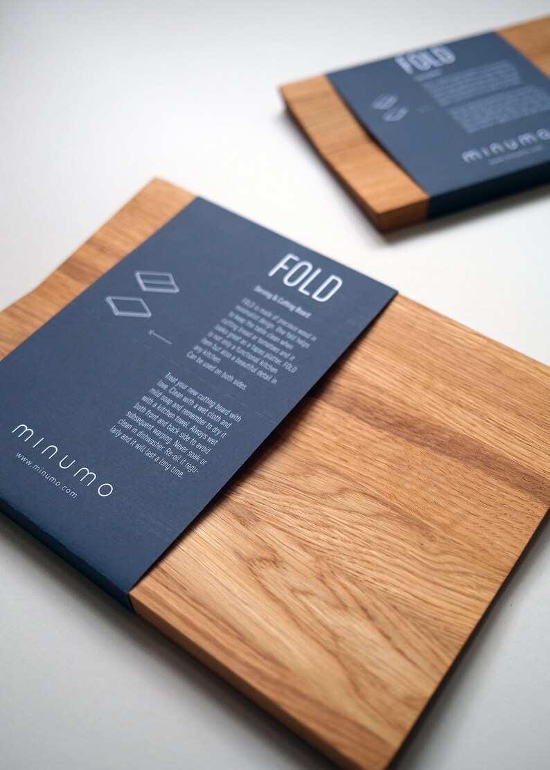 May include: Two rectangular wooden cutting boards with a natural grain finish. Each board features a dark blue paper band wrapped around one end, printed with the white text FOLD, Minumo, and care instructions for the kitchen product.