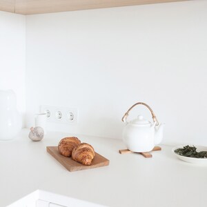 May include: A white ceramic teapot with a curved rattan handle sits on a small wooden trivet. Beside it, two golden-brown croissants rest on a rectangular wooden board. A small white bowl containing loose tea leaves is positioned to the right.