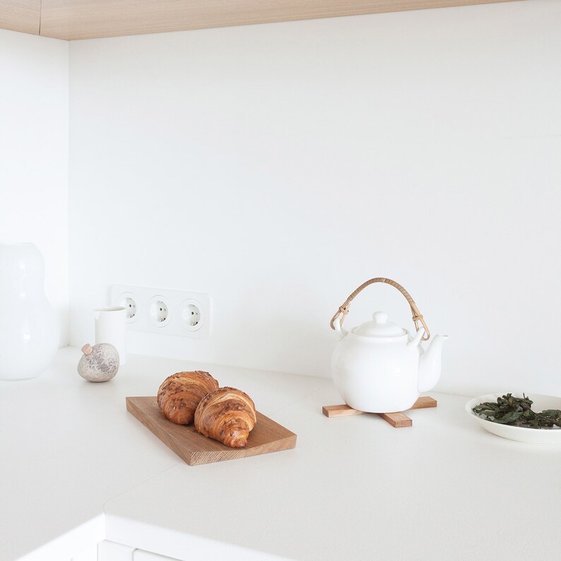 May include: A white ceramic teapot with a curved rattan handle sits on a small wooden trivet. Beside it, two golden-brown croissants rest on a rectangular wooden board. A small white bowl containing loose tea leaves is positioned to the right.