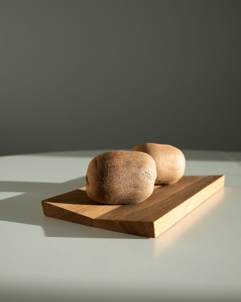 May include: Two whole kiwi fruits with brown, fuzzy skin resting on a rectangular wooden cutting board. The board features a visible wood grain pattern and light brown tones, positioned under direct light creating a sharp shadow.