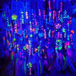May include: A collection of colorful, neon-colored hair extensions with feathers and beads. The extensions are arranged in a vertical row and are glowing under a black light.