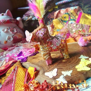 May include: A pink and gold decorative figurine of a horse with a feather and sequins. The figurine is sitting on a wooden surface with a variety of colorful fabric scraps and butterflies.