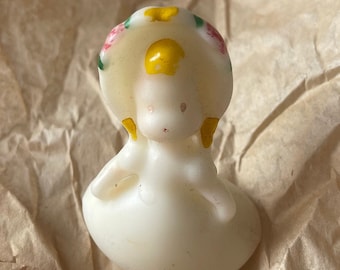 May include: A white candle shaped like a girl wearing a yellow and white hat with pink and green flowers. The candle is sitting on brown paper.