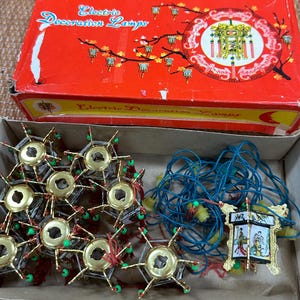 May include: A vintage box of Electric Decoration Lamps. The red box features a circular design with Chinese characters and illustrations. Inside, there are several gold-colored star-shaped lamps with green and red accents, along with a single decorative lantern.