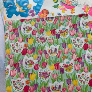 Puede incluir: Un dise&ntilde;o de papel de regalo vintage con un patr&oacute;n repetitivo de tulipes rosas, moradas y amarillas con conejos blancos que sostienen cestas de flores sobre un fondo azul claro.