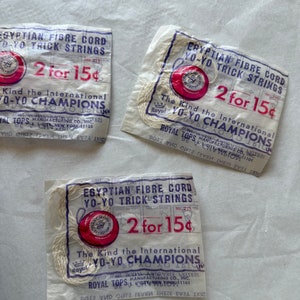 May include: Three white paper packages with red and white yo-yo strings. The packages have the text "Egyptian Fibre Cord Yo-Yo Trick Strings" and "2 for 15¢". The text "The Kind the International Yo-Yo Champions" is also on the packages.