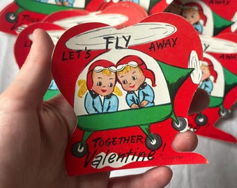 May include: A vintage Valentine's Day card featuring two children in a red and green helicopter. The card reads "Let's Fly Away Together Valentine!"