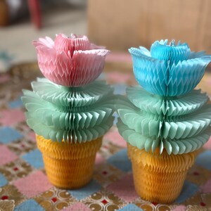 May include: Two decorative paper flower arrangements. Each features a pink or blue flower atop a green, layered base, set in a yellow, honeycomb-style pot. The background is a patterned fabric with pink, blue, and gold elements.