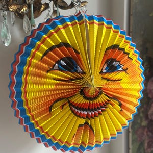 May include: A colorful, round paper lantern with a smiling sun face design. The lantern features a yellow center with orange and red accents, blue and red edges, and a cheerful expression. It is designed to hang and is suitable for festive occasions.