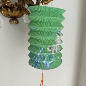 May include: A cylindrical green paper lantern with accordion-style pleats and a hand-painted design featuring blue and white brushstrokes. A small, thin tassel with pink and yellow threads hangs from the bottom center of the lantern.