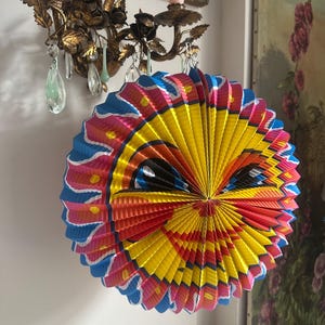 May include: A colorful, round paper lantern with a smiling face design. The lantern features a vibrant palette of yellow, blue, pink, and red. It is hanging on a white wall, adding a cheerful touch to the decor.