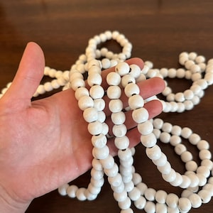 May include: White wooden bead garland. The garland is made of round, painted beads strung together. The beads are held in a hand, with more of the garland visible in the background. The beads are a matte white color.