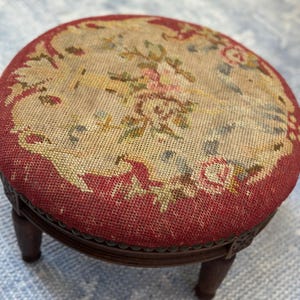 May include: A small, round footstool with a detailed floral tapestry top. The tapestry features pink, green, and yellow flowers on a beige background, framed by a red border. The stool has dark brown, carved wooden legs.