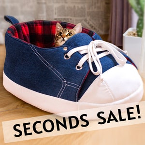May include: A novelty cat bed in the shape of a blue denim trainer, with white laces and toe cap. The interior is lined with red and black plaid fabric. A cat is visible inside. The text "SECONDS SALE!" is displayed.