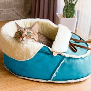 May include: A cat rests in a blue and cream-colored pet bed shaped like a shoe. The bed has a soft, plush interior and brown leather-like laces. The cat has green eyes and brown and tan fur.