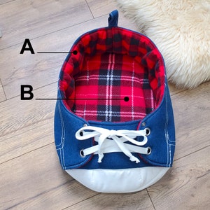 May include: A blue denim-style pet bed shaped like a sneaker. The interior features a red and black plaid pattern. White laces and a white sole complete the design. A loop is attached at the top for easy carrying.