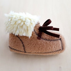 May include: A single baby bootie in a light brown color with a cream-colored, fluffy trim. The bootie has a dark brown bow and is stitched with black thread. The overall design suggests a cozy, handmade item.