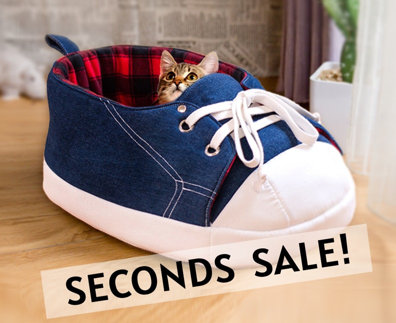 May include: A blue denim cat bed shaped like a high-top sneaker with white laces and a white toe cap. The interior is lined with red and black plaid fabric. A cat peeks out from inside. The text "SECONDS SALE!" is displayed.