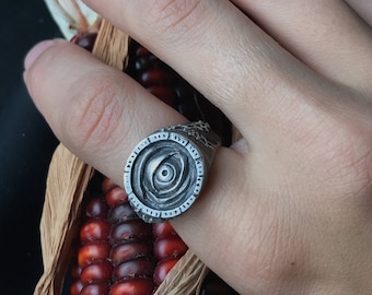 May include: A silver ring with a circular design featuring a raised eye in the centre. The ring is worn on a finger with a small tattoo.