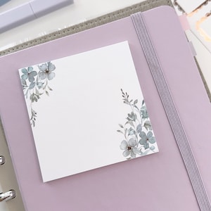 May include: A white sticky note with a floral design in shades of blue and green. The note is attached to a light purple planner with a gray elastic band.