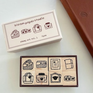 May include: A set of eight wooden stamps in a dark brown box, accompanied by a white cardboard sleeve labeled bloompaperstudio stamp set vol. 1 8pcs. The stamps feature black line art illustrations of various bags, stationery, and office items.