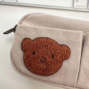 May include: A beige pencil case with a brown teddy bear applique. The pencil case has a zippered closure and a small pocket on the front. The teddy bear applique is made of a textured, brown material.
