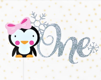 May include: A cake topper featuring a cartoon penguin with a pink bow, bright pink cheeks, and an orange beak. Next to the penguin is the word One in silver glitter script, accented with two silver glitter snowflakes on a white stick base.