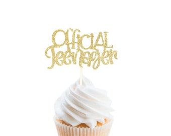 May include: A gold glitter cupcake topper featuring the text Official Teenager in a cursive and print font combination, attached to a thin wooden pick and inserted into a white frosted cupcake with a light tan paper liner.