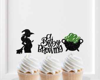 May include: Three cupcake toppers featuring black glitter silhouettes of a pregnant witch with a broom, the text "a Baby is Brewing" in black glitter, and a black glitter cauldron with green glitter bubbles, each mounted on a small white stick.