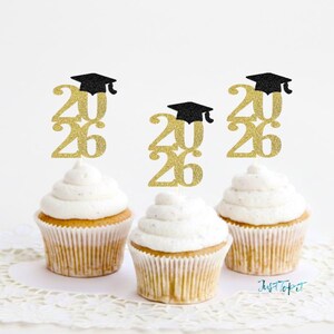 May include: Three cupcakes topped with gold glittery 2026 cake toppers featuring a black graduation cap. Each topper is positioned vertically in the center of the white frosting swirl on top of the light brown baked cupcake base.