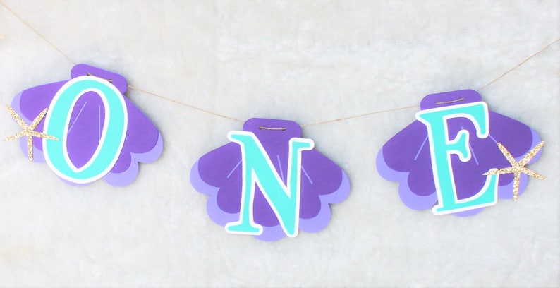 May include: A decorative banner featuring three purple seashell-shaped cutouts strung together on a thin cord. Each shell displays a large, bright turquoise letter spelling out the word ONE, accented with small, gold glittery starfish.