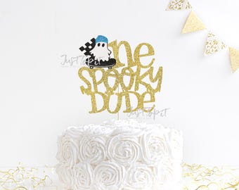 May include: A gold glitter cake topper featuring the text "one SPOOKY DUDE" with a graphic of a white ghost wearing a blue hat on a black skateboard. The topper is positioned above a white cake with a swirled frosting texture and gold accents.