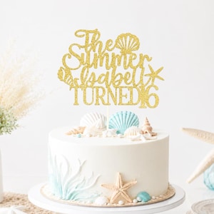 May include: A gold glitter cake topper featuring the text The Summer Isabell TURNED 16 with seashell and starfish accents. The topper is positioned on a white cake decorated with light blue and white seashells and a starfish on the side.
