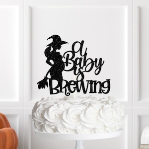 May include: Black glitter cake topper featuring a silhouette of a pregnant witch holding a broom with the text "A Baby is Brewing".