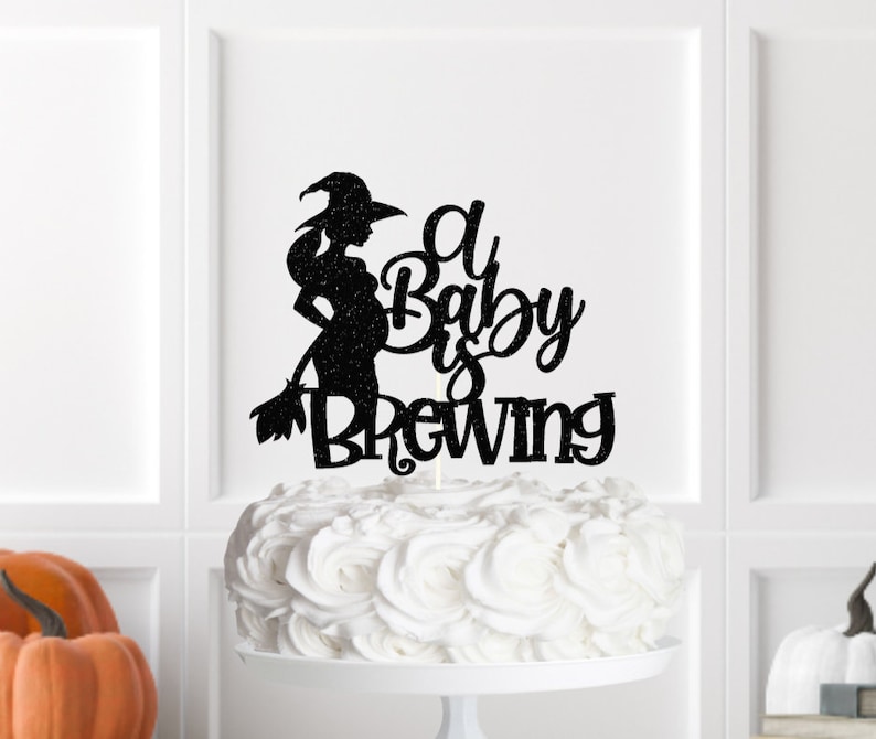 May include: Black glitter cake topper featuring a silhouette of a pregnant witch holding a broom with the text "A Baby is Brewing".