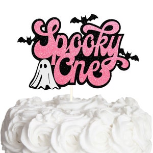 May include: A cake topper featuring the text Spooky One in pink glittery script with a black glittery outline. The design includes a white ghost figure on the left and three black bat silhouettes positioned around the lettering, all mounted on a stick.