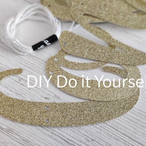 May include: Gold glitter letters for a DIY banner, with holes punched for stringing. The letters are on a white wooden surface. The text "DIY Do it Yourself!" is visible in the image.