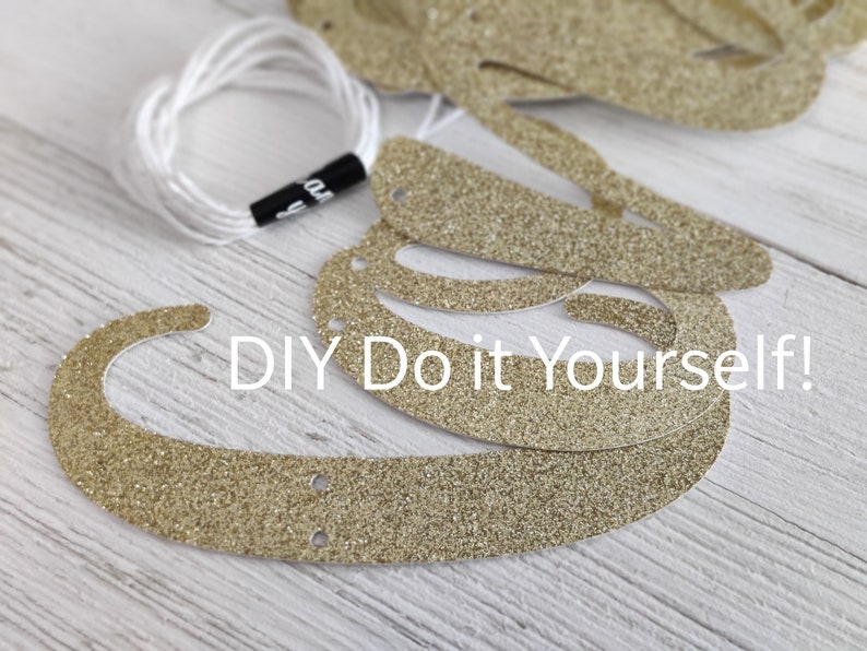 May include: Gold glitter letters for a DIY banner, with holes punched for stringing. The letters are on a white wooden surface. The text "DIY Do it Yourself!" is visible in the image.