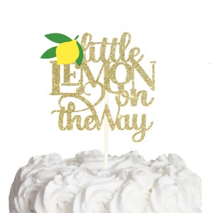 Op de afbeelding: Een gouden glitter taarttopper met de tekst Little Lemon on the Way in een combinatie van script- en blokletters. Een gele citroenafbeelding met twee groene bladeren bevindt zich boven de tekst. De topper zit vast aan een dun wit stokje.