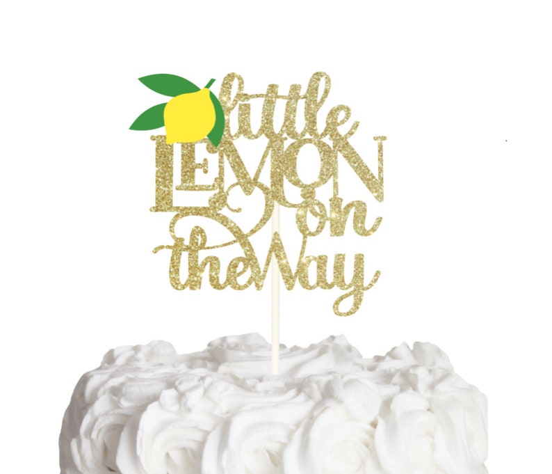 Op de afbeelding: Een gouden glitter taarttopper met de tekst Little Lemon on the Way in een combinatie van script- en blokletters. Een gele citroenafbeelding met twee groene bladeren bevindt zich boven de tekst. De topper zit vast aan een dun wit stokje.