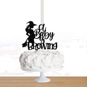 May include: Black glitter cake topper featuring a silhouette of a pregnant witch holding a broom with the text "A Baby is Brewing".