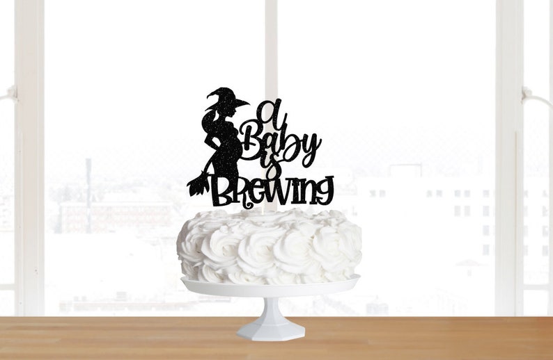 May include: Black glitter cake topper featuring a silhouette of a pregnant witch holding a broom with the text "A Baby is Brewing".