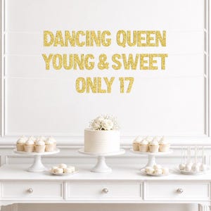 May include: A gold glitter banner with the text DANCING QUEEN YOUNG & SWEET ONLY 17 suspended on white strings. Below the banner is a white table holding a white cake topped with white flowers, white cupcakes on stands, and white cake pops.