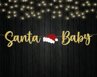 May include: A decorative banner featuring the gold glitter text Santa Baby with a red and white Santa hat positioned between the two words. The letters are connected by a thin string against a dark wood background with soft fairy lights.