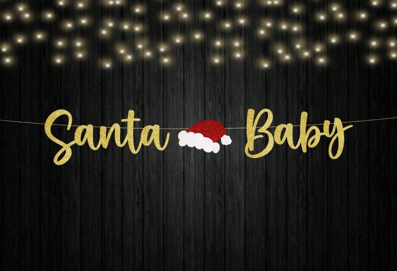 May include: A decorative banner featuring the gold glitter text Santa Baby with a red and white Santa hat positioned between the two words. The letters are connected by a thin string against a dark wood background with soft fairy lights.