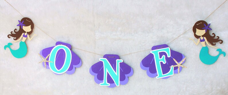 May include: A decorative banner featuring two mermaid cutouts with brown hair and teal tails, flanking three purple seashell cutouts. Each shell displays a large light blue letter, spelling out the word ONE, accented with small gold glitter starfish.