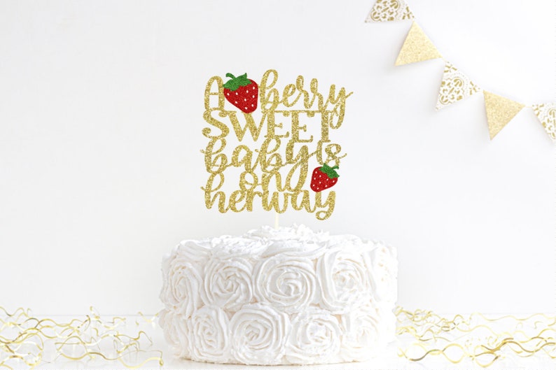 May include: Gold glitter cake topper with the text "A berry sweet baby is on her way" and two red strawberries.