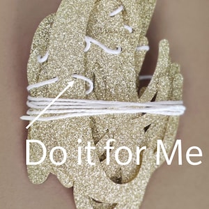 May include: Gold glitter letters spelling "Do it for Me" strung together with white string. The letters are layered on top of each other.